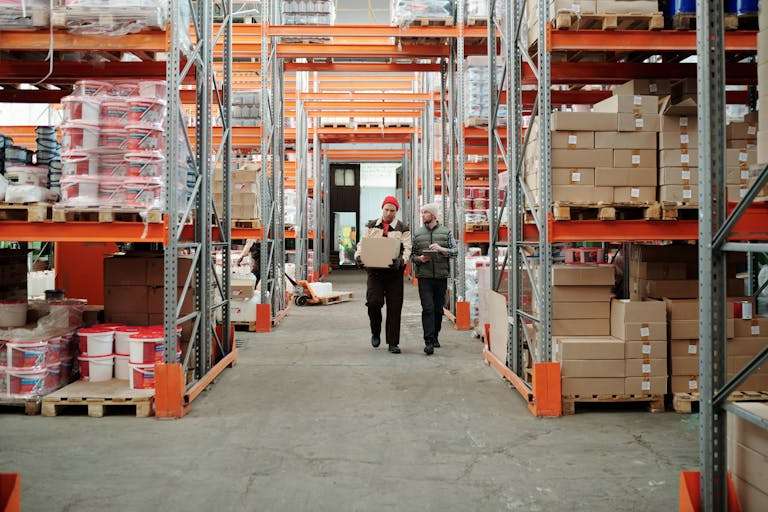 Two workers handle a package in a spacious warehouse surrounded by shelves stocked with boxes and products.